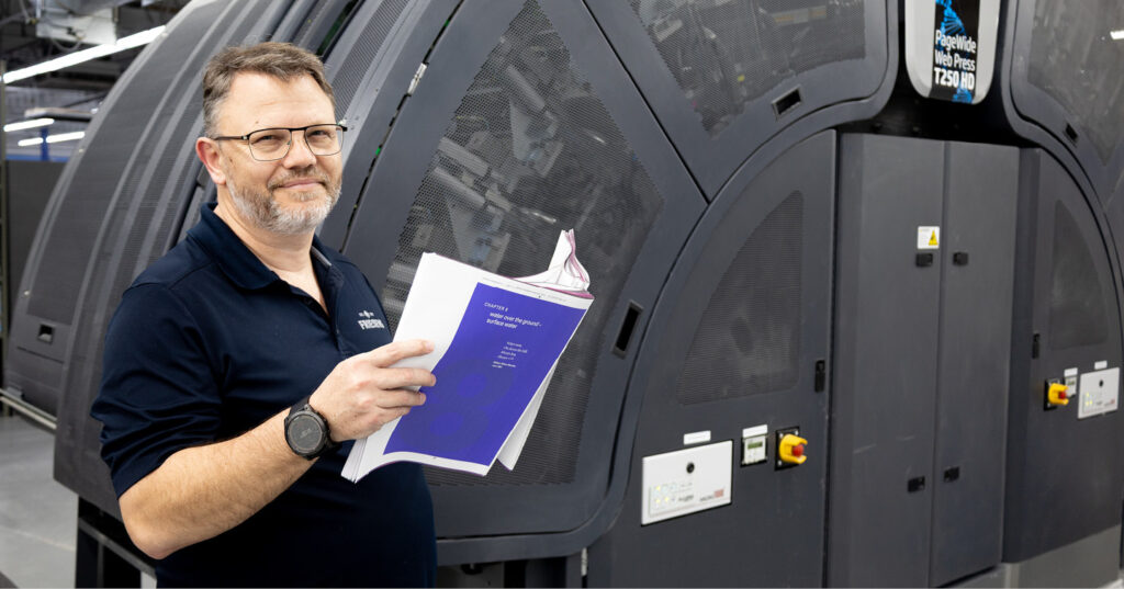 Person holding printed pages beside a large inkjet web press in a printing facility.