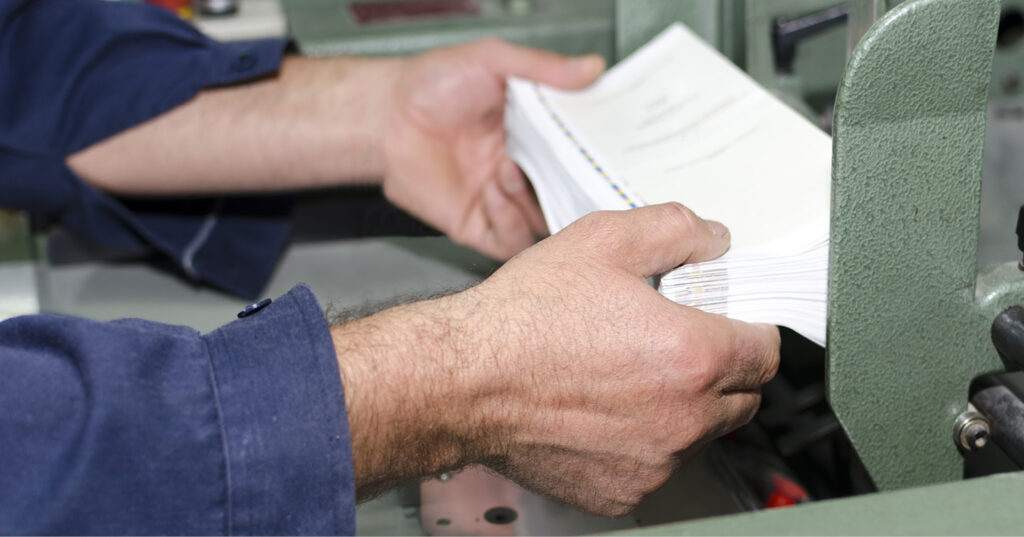 Close-up of hands aligning a stack of printed pages at an industrial printing machine.