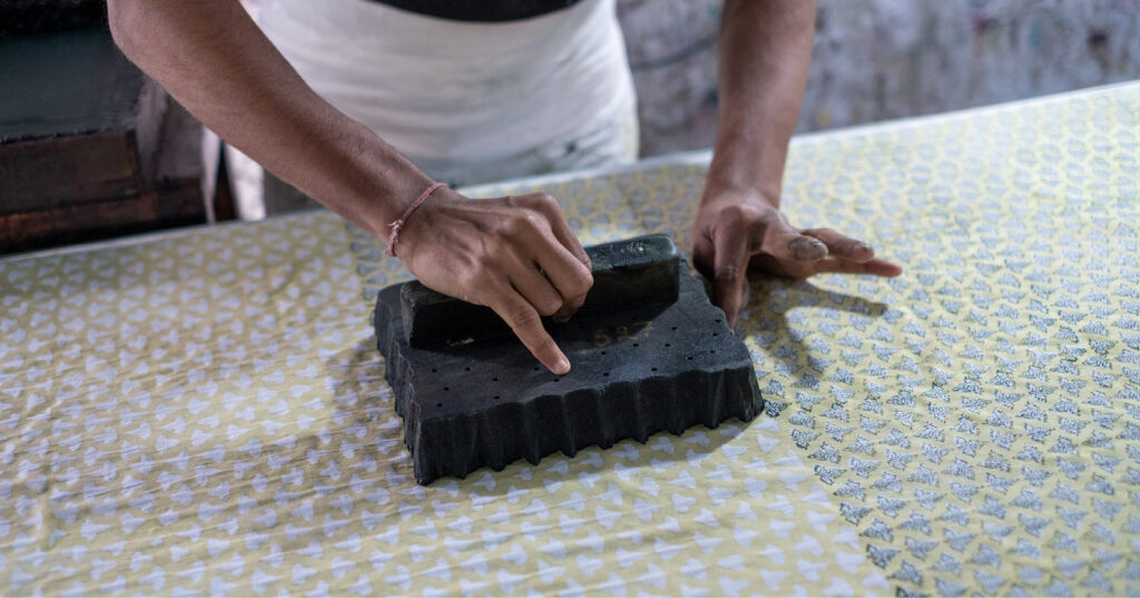 Hands pressing a carved printing block onto patterned fabric, demonstrating traditional textile block printing.