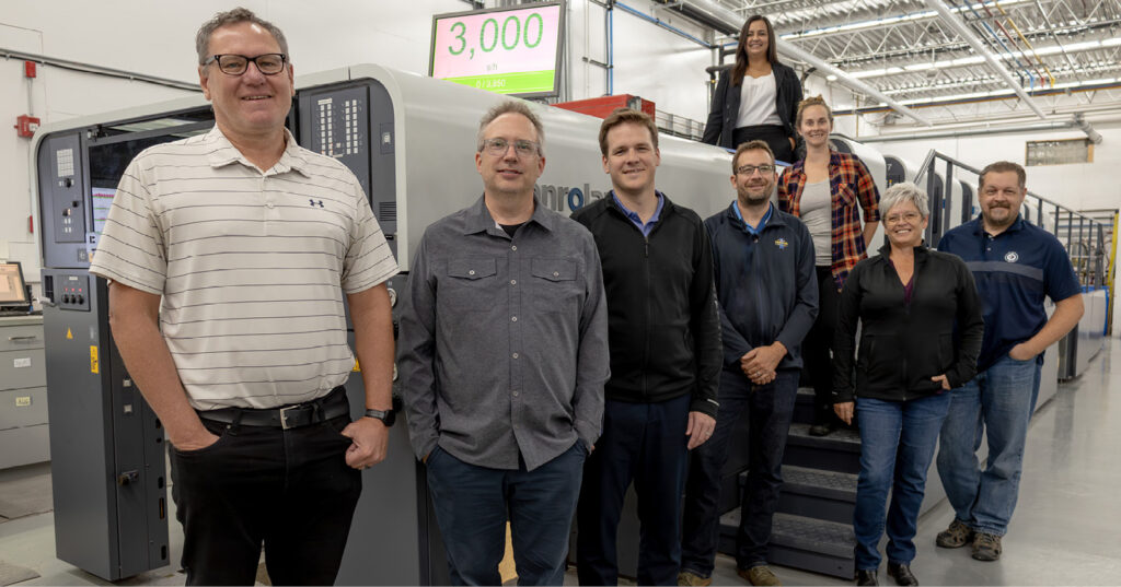 Employees stand together next to an offset printing press on a production floor.