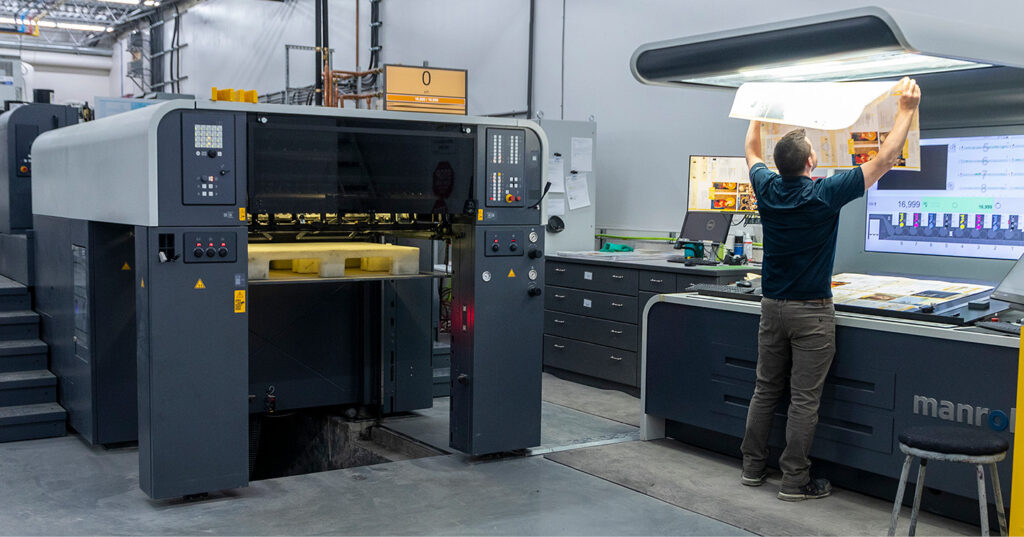 Press operator inspects printed sheets under light beside large offset printing press and control screens