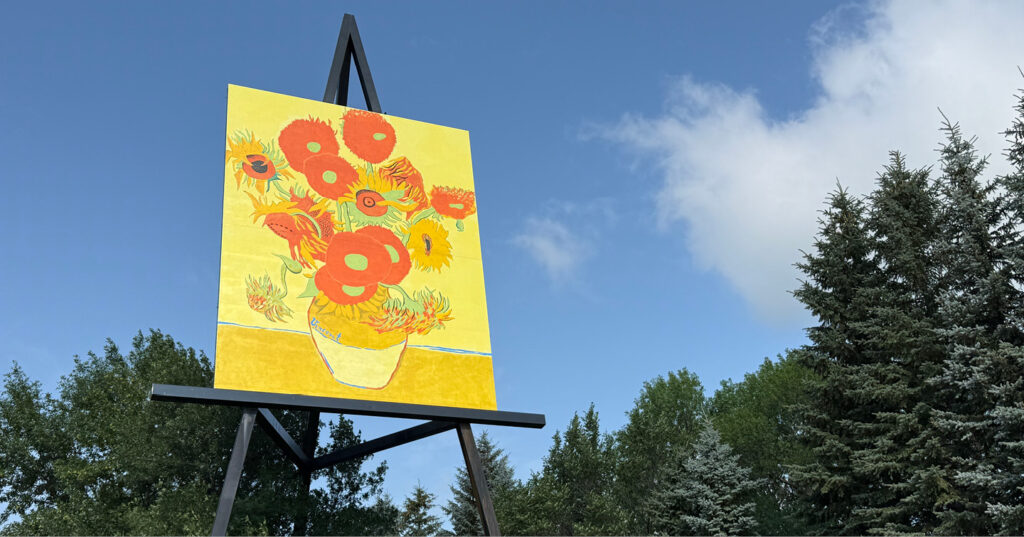 Large outdoor easel displays a sunflower painting against a blue sky, surrounded by trees and greenery.