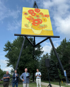 People gather beneath a giant outdoor easel displaying a sunflower painting during a community event.