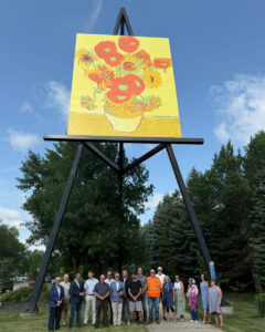Group photo beneath a giant outdoor easel displaying a sunflower painting, surrounded by trees and blue sky.