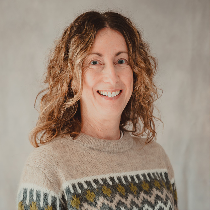Professional headshot of a person with curly hair wearing a patterned knit sweater against a neutral studio background.