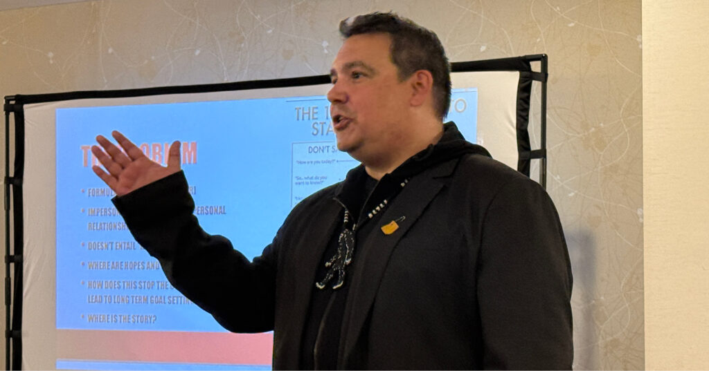 Niigaan Sinclair speaks to the Friesens team during a presentation, standing in front of a screen displaying slides.