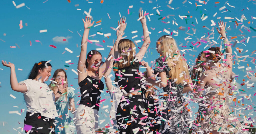 Students celebrating outdoors as colorful confetti falls around them under a bright blue sky.