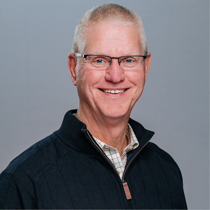 Professional headshot of a person wearing glasses and a dark quarter-zip sweater against a gray background.