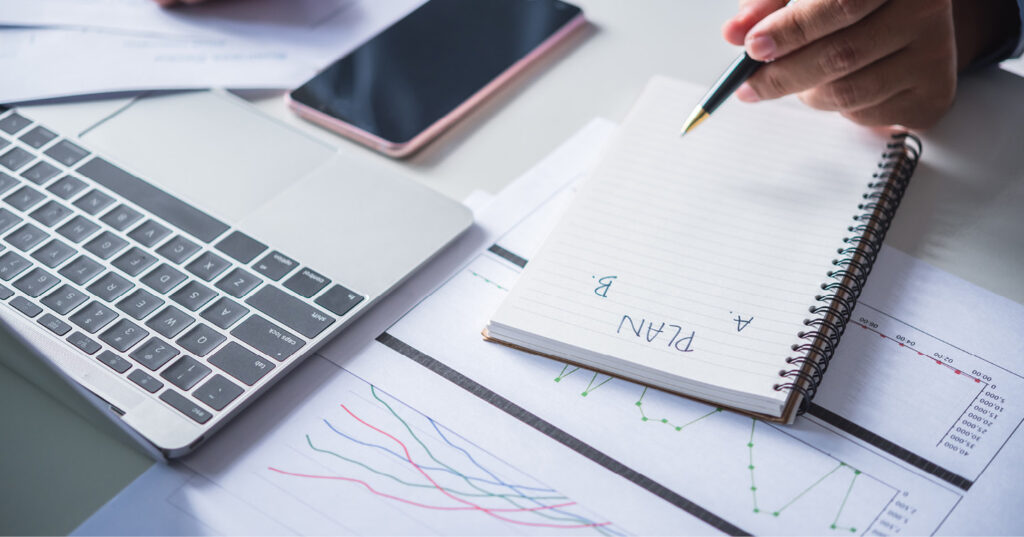 Hand writing in a notebook beside a laptop, phone, and charts during business planning and data analysis.