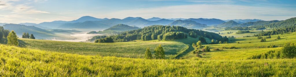 Rolling green hills and forested mountains stretch into the distance beneath a clear blue morning sky.
