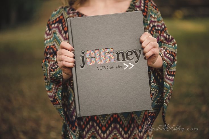 Person holding a gray yearbook with foil-accented “Journey” cover design, photographed outdoors with blurred background.