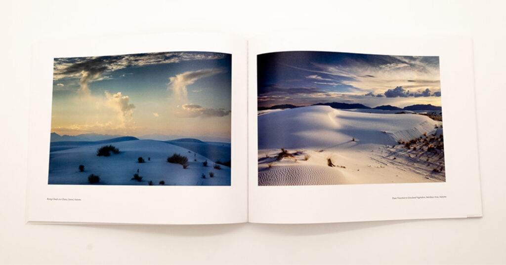 Open coffee table book displaying two full-page photographs of white sand dunes beneath dramatic skies and distant mountains.