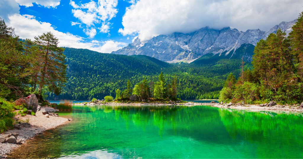 Crystal-clear turquoise lake surrounded by evergreen forest and rocky mountains, with clouds drifting over the peaks.