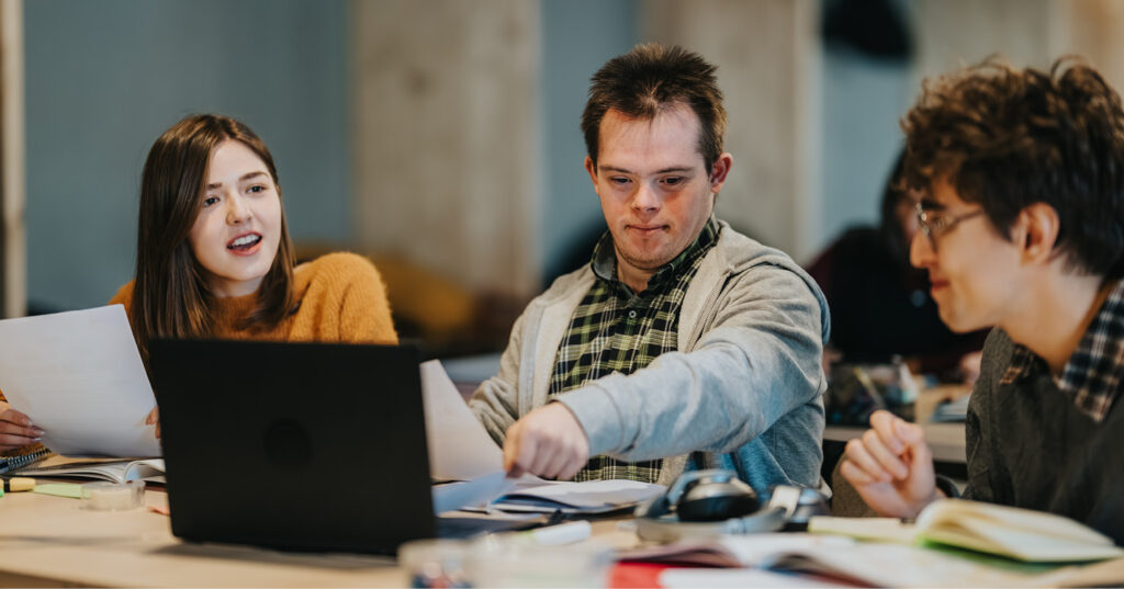 Three students collaborate at a table with a laptop, notebooks, and papers during a group study session.